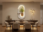 A modern dining room setup featuring a dark wood dining table surrounded by six upholstered chairs, with a decorative sideboard against a beige wall. The focal point is a large, oval-shaped white wall mirror with an integrated vertical shelf holding a potted bamboo plant, illuminated by warm lighting from three hanging pendant lights.