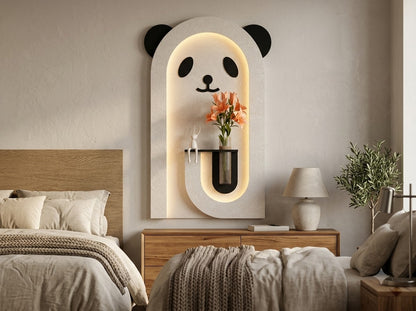 A modern wall-mounted panda-face light fixture in a bedroom setting, featuring a white arched frame with black panda ears, eyes, and nose, and warm LED backlighting. A small vase with orange flowers is mounted in the center of the fixture.