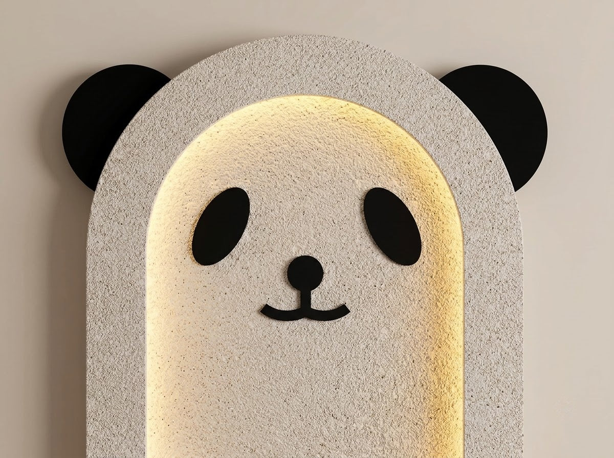 A modern wall sconce light fixture shaped like a panda face, featuring a textured light gray stone-like frame with black ears, eyes, nose, and mouth, illuminated from within by a warm yellow light against a beige wall.